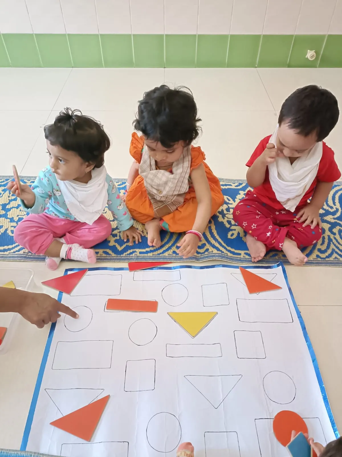 Toddlers matching wooden shapes on an activity mat with a caretaker guiding them